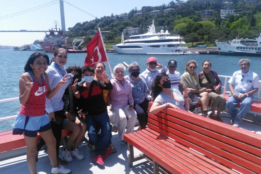 WhatsApp Image 2022-12-05 at 12.48.07 (2) Un numeroso grupo de turistas en un paseo en barco por el Bósforo; los pasajeros posan frente a la bandera turca en la cubierta del barco, con el puente del Bósforo, yates de lujo y la costa verde de fondo.