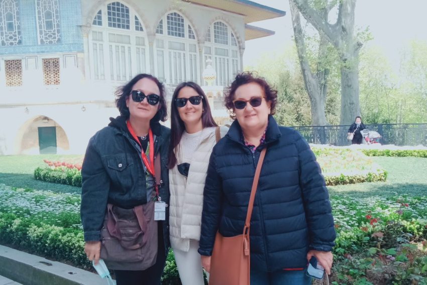 Tres turistas sonrientes posan una al lado de la otra en el jardín frente al Pabellón de Bagdad del Museo del Palacio de Topkapi; al fondo, se aprecia el exterior de azulejos del histórico pabellón y árboles centenarios.