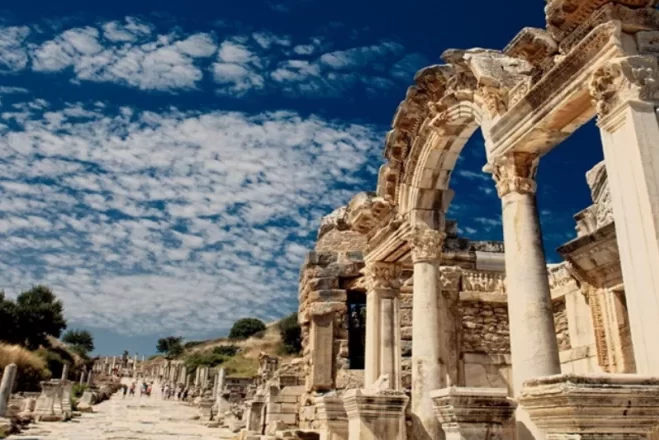 La estructura de arcos y columnas de mármol del histórico Templo de Adriano en la calle Curetes, en la antigua ciudad de Éfeso; al fondo, un paseo de bloques de mármol y un cielo azul parcialmente nublado.