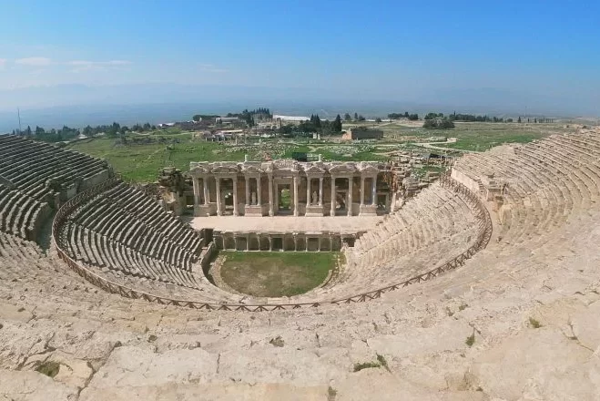 Vista panorámica desde los escalones superiores del antiguo teatro de Pamukkale en Hierápolis; se aprecia el edificio del escenario con columnas, bien conservado, las zonas de asientos semicirculares de piedra y la llanura que se extiende al fondo.