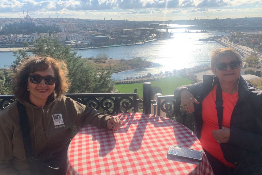 Dos mujeres con gafas de sol están sentadas en una mesa cubierta con un mantel a cuadros en la terraza de una cafetería con vistas al Cuerno de Oro en Estambul. Detrás de ellas, se divisa una vista panorámica de la ciudad, que abarca puentes, mezquitas y el Cuerno de Oro.