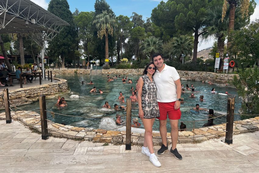 WhatsApp Image 2024-12-31 at 09.06.02 "Una pareja sonríe y posa frente a las aguas turquesas de la histórica Piscina Antigua en Pamukkale."