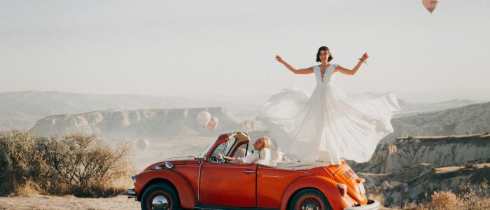 Al amanecer en Capadocia, una novia con su vestido de boda ondea al viento sobre un clásico coche rojo, mientras globos aerostáticos flotan en el cielo.